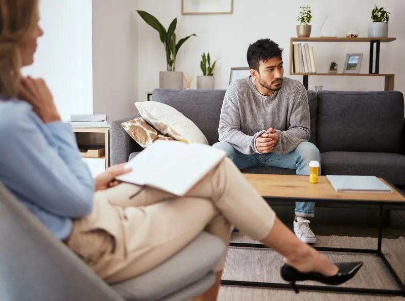 A psychotherapist in Brooklyn is listening attentively as a client discusses their mental health concerns in a comfortable therapy setting. The client looks reflective while sitting on the couch.