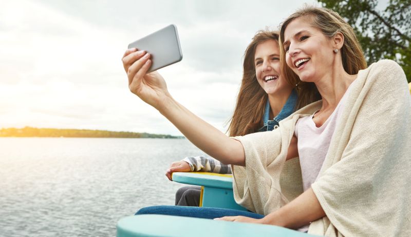 Two women sharing a joyful moment together by the lake, smiling while taking a selfie. Therapists for postpartum depression offer essential emotional support during life changes like motherhood