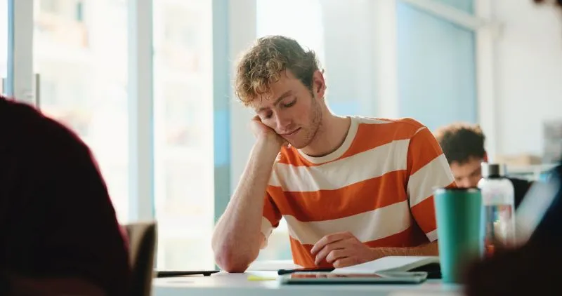 A student in a classroom appearing distracted, resting his head on his hand while working on his tasks. An ADHD therapist could help address his focus and engagement issues.