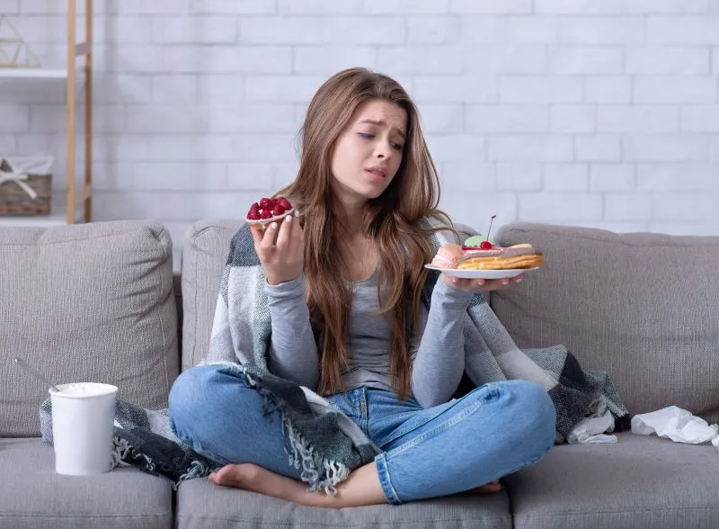 A teenage girl sitting on the couch, holding a bowl of grapes in one hand and a plate of pancakes in the other, displaying a conflicted expression. This reflects the internal struggle that often accompanies binge eating disorder, where individuals may feel conflicted between healthy and unhealthy food choices.