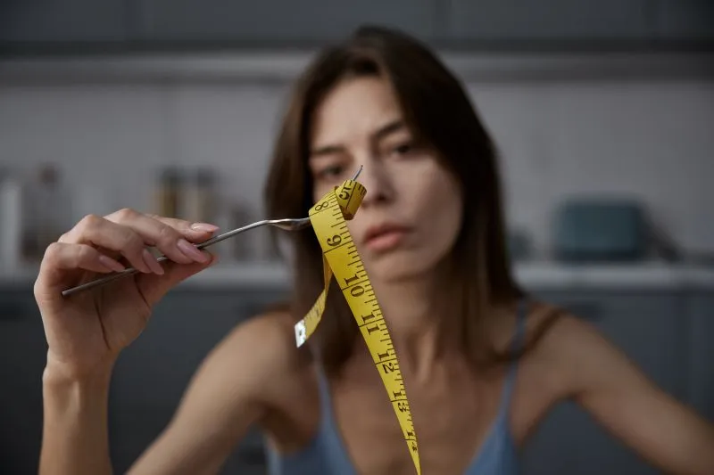 A woman holding a measuring tape next to her food, possibly overthinking her food intake and body image, which is common in those struggling with binge eating disorder, where there is a preoccupation with weight and food control.