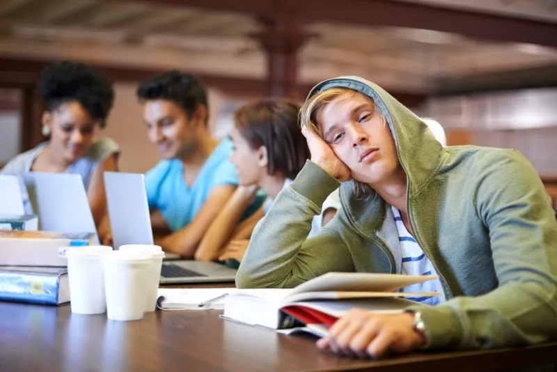 A teenager in a study group, appearing disengaged and exhausted while others focus on their laptops. ADHD specialists can help improve concentration and engagement for individuals with attention difficulties.