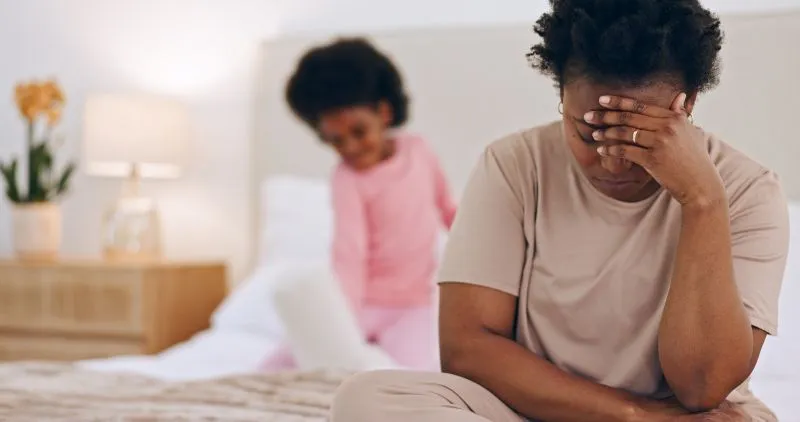 A mother sitting on a bed, looking stressed, while her child plays in the background. Therapists specializing in ADHD can support parents in managing their child’s symptoms and improving family dynamics.