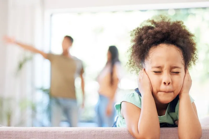 A young girl covering her ears with her hands, distressed by the tension between her parents in the background. This visual reflects the emotional impact of childhood trauma and its potential symptoms in adulthood