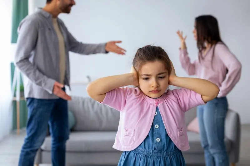 A young girl looks worried and covers her ears as her parents argue behind her. This scene represents how childhood trauma, such as witnessing conflict, can lead to emotional challenges in adulthood.