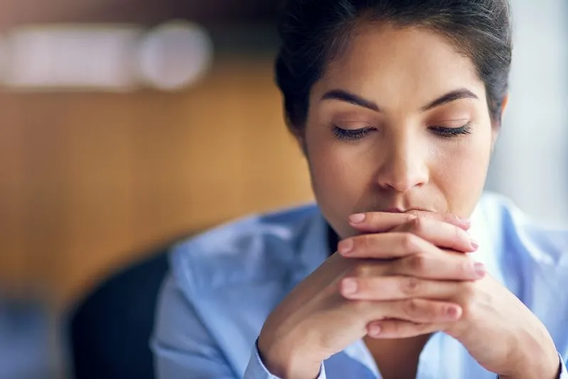 A woman in a professional setting looking stressed and contemplative, with her hands clasped together. This image highlights how individuals with ADHD may experience mental health struggles, such as anxiety and depression.