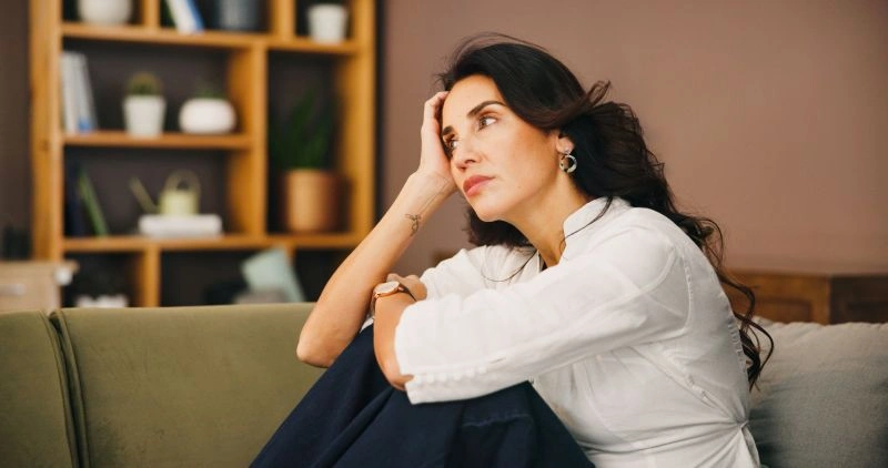 A woman sitting on a couch, gazing thoughtfully into the distance while holding her head. This moment of reflection captures how the challenges of ADHD can contribute to feelings of anxiety and depression.