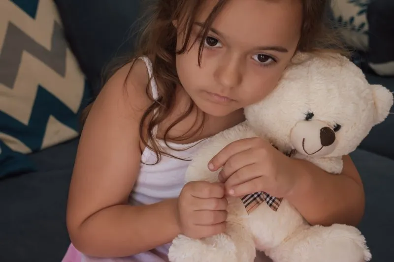 A young girl holding a teddy bear with a sad expression, reflecting the emotional distress that may stem from childhood trauma and the journey to heal from it.