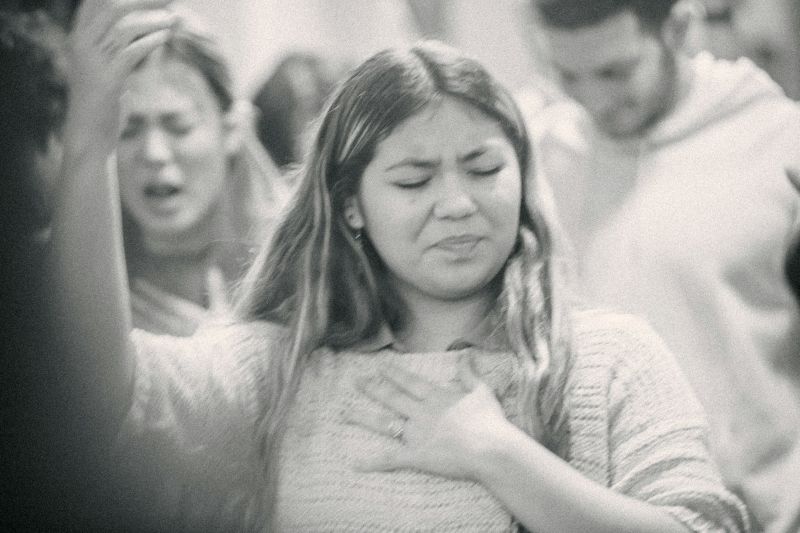 grayscale image of a woman in a crowded setting with eyes closed and hand on her chest, appearing emotionally overwhelmed in a social environment.