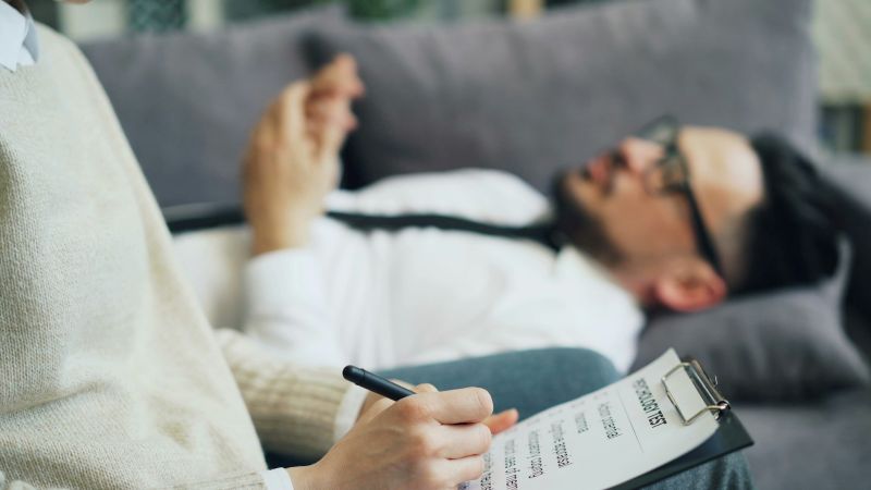 mental health professional taking notes during a consultation while a patient lies on a couch describing symptoms during evaluation.