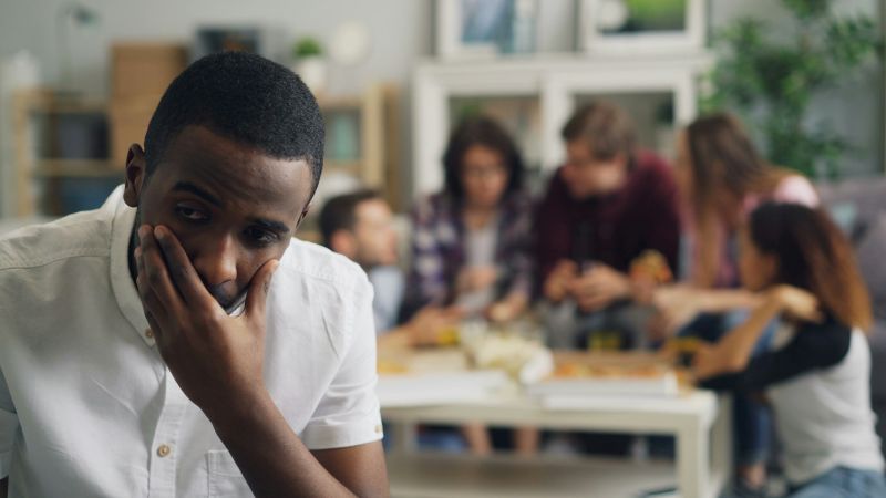 man sitting alone in a room with a group of people in the background, appearing withdrawn and socially anxious