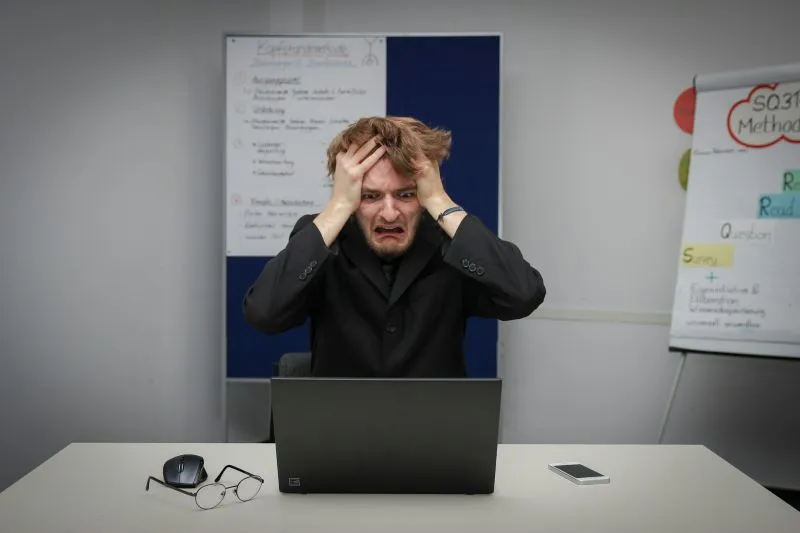 Emotional volatility shown by a distressed man holding his head while sitting at a laptop in an office.